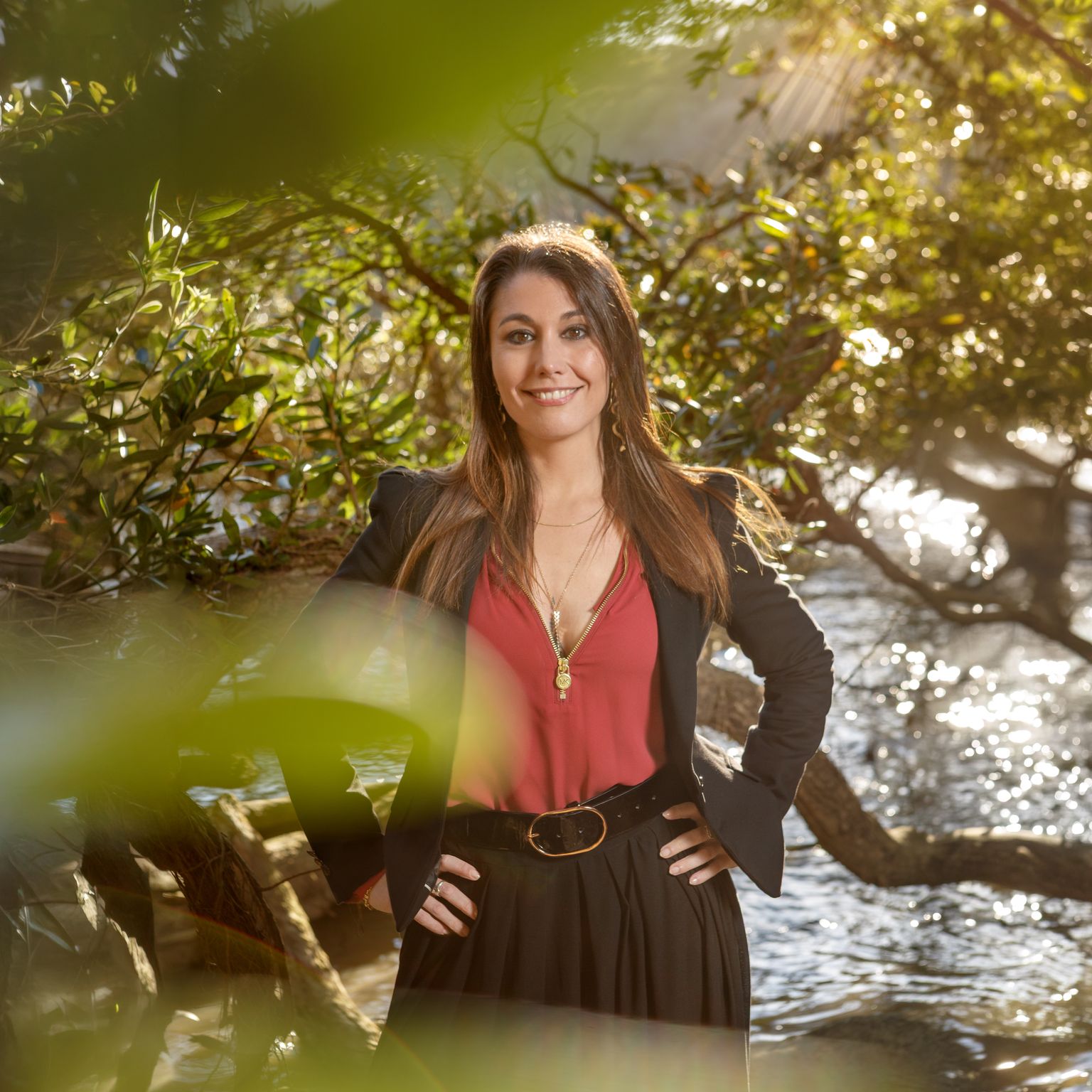 Alma in a dark blazer standing among coastal trees by the water